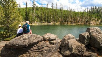 Two Hikers Take Lunch On A Rock While Overlooking A Small Lake In Yellowstone National Park